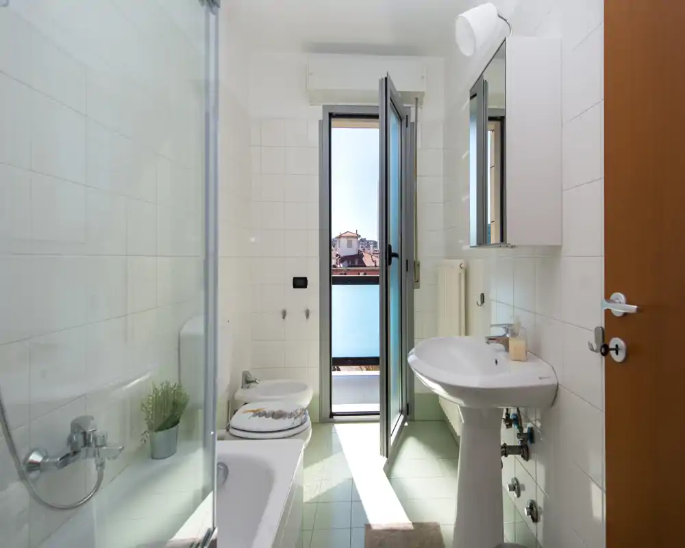 Bagno luminoso e moderno con vasca, bidet, lavabo e porta a vetri che si apre su un piccolo balcone con luce naturale.