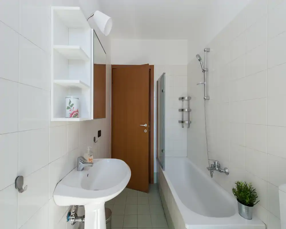 Bagno pulito e luminoso con vasca, doccia, lavabo e armadietto con specchio a parete. Piastrelle bianche e porta in legno offrono un aspetto fresco e minimale.
