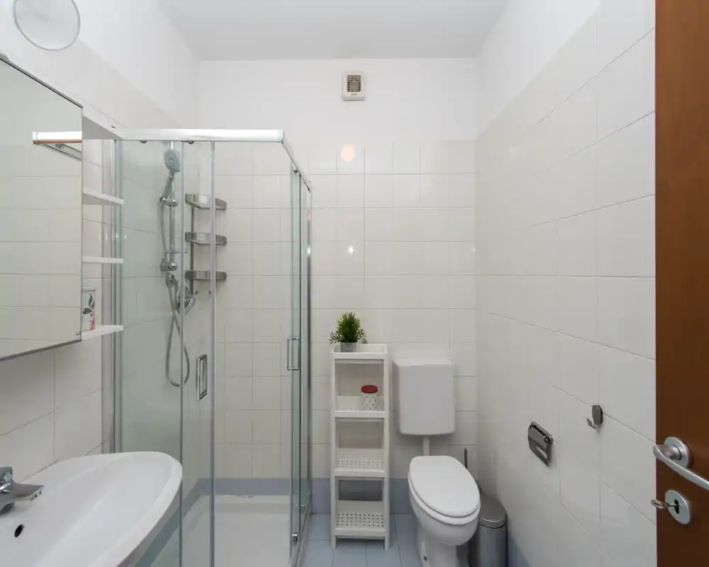 Bagno compatto e ben illuminato con box doccia in vetro, cassetta del WC a parete, lavabo con specchiera e piccolo scaffale per riporre oggetti.