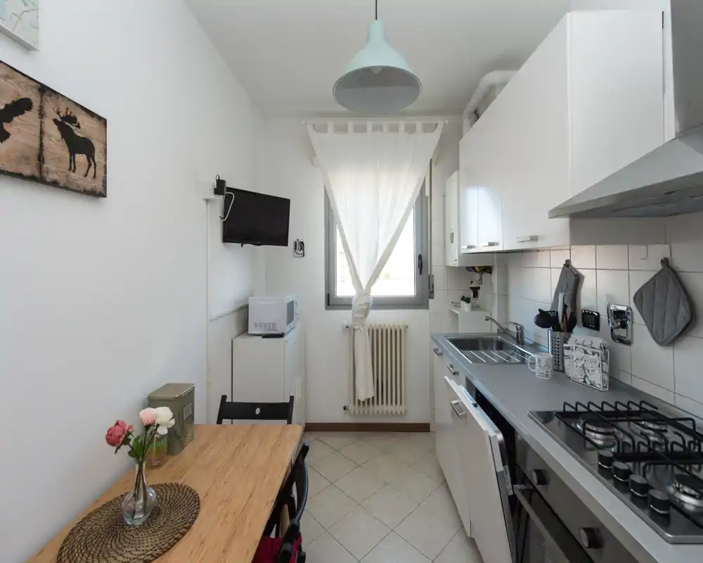 Cucina compatta e ordinata con elettrodomestici moderni, piano cottura a gas, lavello e un piccolo tavolo in legno vicino alla finestra che crea uno spazio luminoso e funzionale.