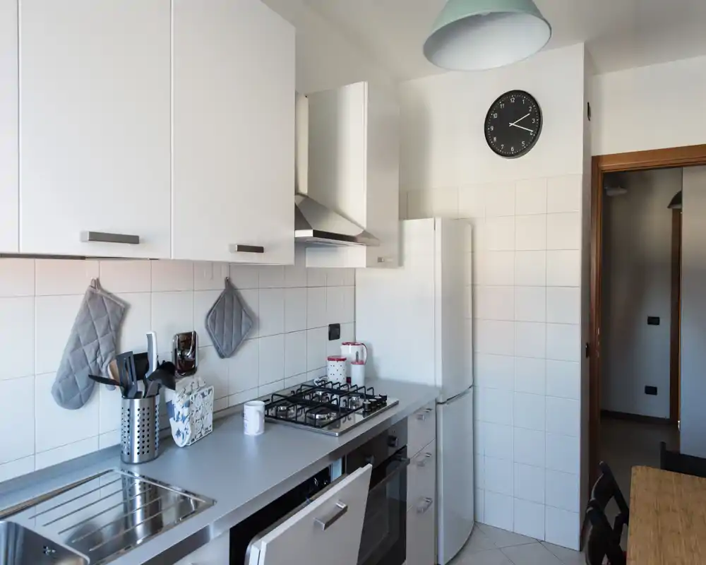 Cucina moderna e luminosa con piano cottura a gas, forno incassato, lavabo in acciaio e pensili bianchi. Accessori ordinati sul piano e un piccolo tavolo rendono l'ambiente funzionale e accogliente.