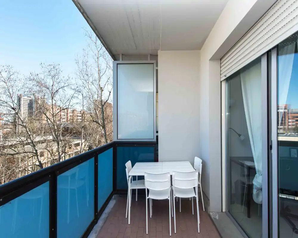 Balcone luminoso con tavolo e quattro sedie, parapetto in vetro e vista sulla città — ideale per pranzi all'aperto o relax.