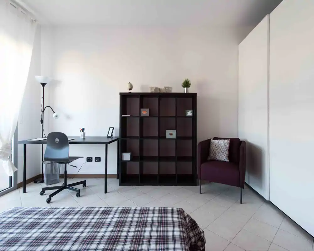 Camera luminosa e minimalista con letto ben visibile in primo piano, scrivania vicino alla finestra, libreria a cubi e una poltrona—ideale per una o due persone.