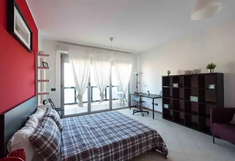 Bright, modern bedroom with a large double bed, patterned duvet, workspace by the window and a shelving unit. Floor-to-ceiling doors open to a balcony bringing abundant natural light.
