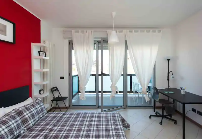 Bright, modern bedroom with a double bed dressed in patterned bedding, a red accent wall, desk workspace and large glass doors opening to a balcony — ideal for a comfortable stay with natural light.