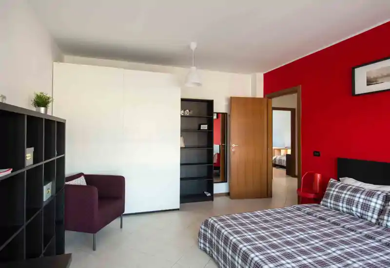 Bright, modern bedroom with a large double bed dressed in checked bedding, a bold red accent wall, wardrobe and shelving; comfortable armchair and simple pendant light complete the space.