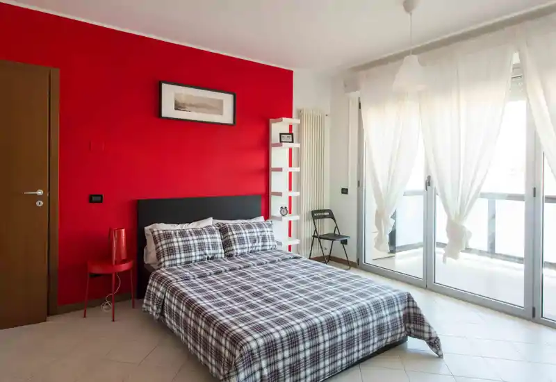 Bright, modern bedroom with a double bed against a bold red accent wall, plaid bedding, and large sliding windows that fill the room with natural light.