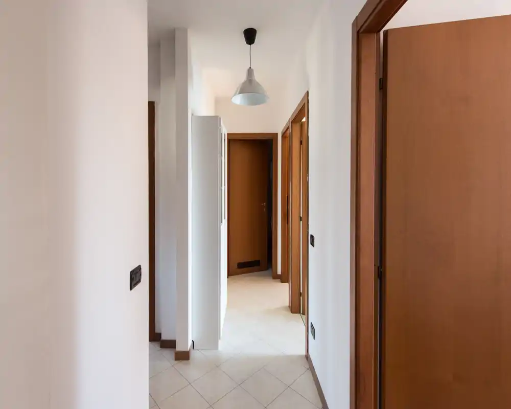 Ingresso/corridoio luminoso e minimale con pavimento piastrellato, pareti bianche e porte in legno; lampada a sospensione semplice e ottimo spazio di passaggio.