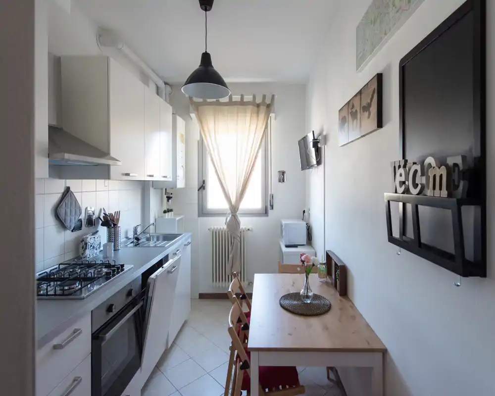 Cucina compatta e luminosa con mobili bianchi moderni, piano cottura a gas, lavello e un piccolo tavolo in legno vicino alla finestra — perfetta per un appartamento urbano accogliente.