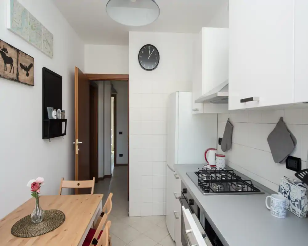 Cucina luminosa e compatta con mobili bianchi moderni, piano cottura a gas, frigorifero e un piccolo tavolo in legno. Arredo neutro e accessori semplici creano un ambiente pulito e accogliente, ideale per affitti brevi.