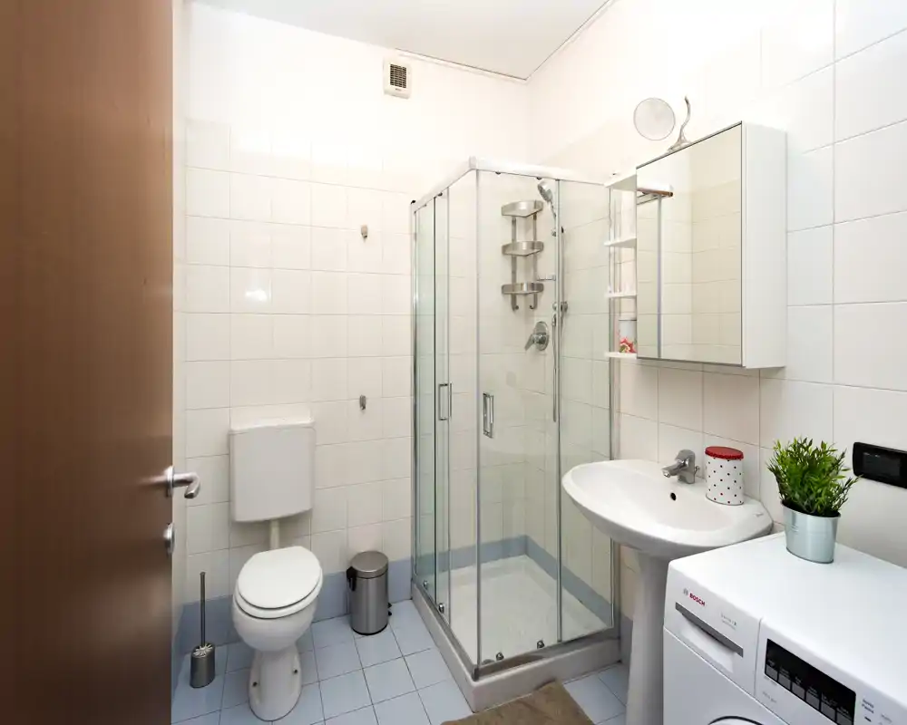 Bagno pulito e ben illuminato con box doccia in vetro, lavabo con mobiletto specchio, WC e lavatrice — disposizione pratica e funzionale.
