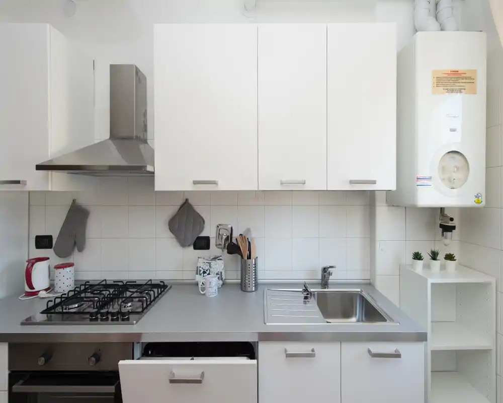 Cucina moderna e luminosa con piano cottura a gas, lavello in acciaio e pensili bianchi. Piano di lavoro ordinato con piccoli oggetti decorativi e utensili pratici.