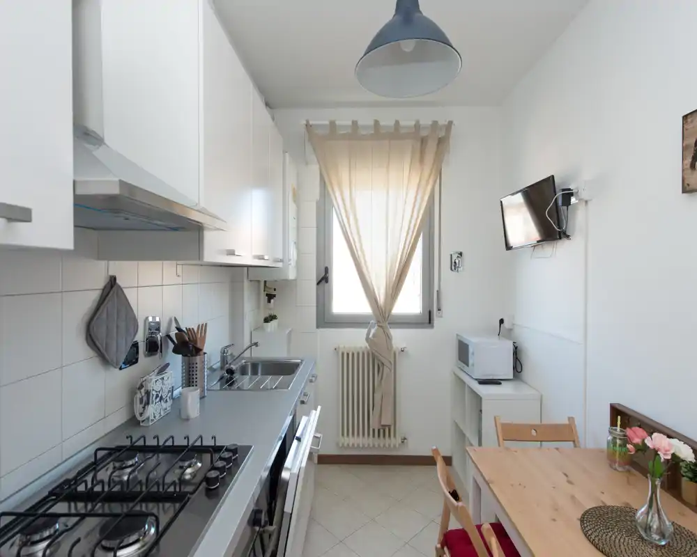 Cucina compatta e luminosa con elettrodomestici moderni, piano cottura a gas, lavello in acciaio e un piccolo tavolo in legno vicino alla finestra.