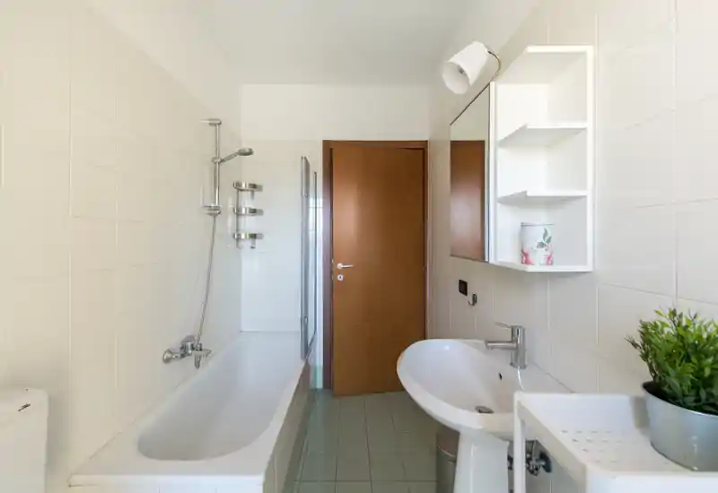 Bright, clean bathroom with a bathtub, wall-mounted shower, sink, and shelving. Neutral tiles and simple fixtures create a fresh, functional space.