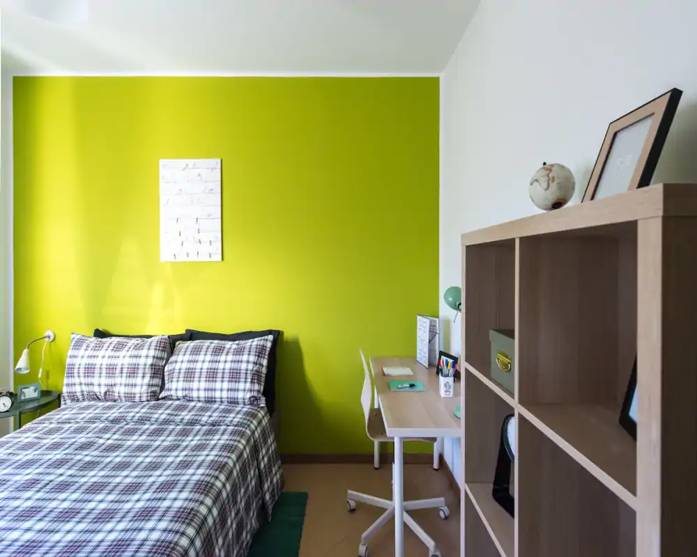 Dormitorio moderno y luminoso con cama doble frente a una pared verde intensa, ropa de cama a cuadros, lámpara de mesita y un pequeño escritorio — ideal para una estancia acogedora.