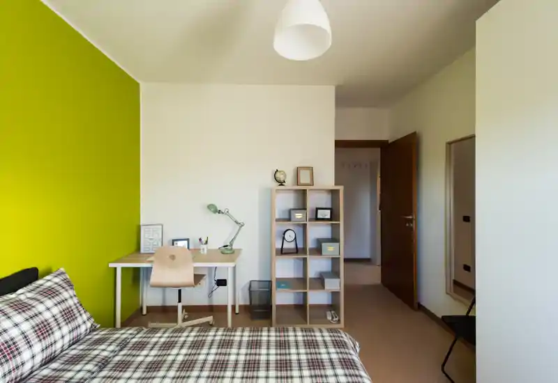 Dormitorio moderno y luminoso con cama estampada, escritorio y estantería frente a una pared verde llamativa — líneas limpias y distribución funcional, ideal para estudiantes o profesionales solteros.