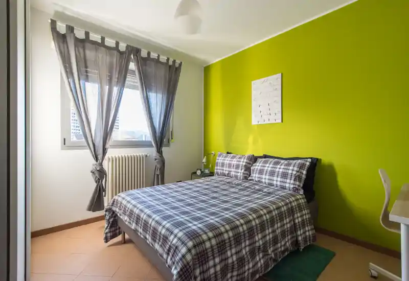 Dormitorio luminoso y ordenado con cama doble vestida con ropa de cuadros, una pared verde llamativa y luz natural de una gran ventana; espacio compacto y bien decorado ideal para parejas o viajeros solos.