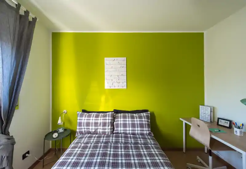 Dormitorio luminoso y bien presentado con cama doble frente a una pared verde llamativa, ropa de cama a cuadros, mesita y un pequeño espacio de trabajo.