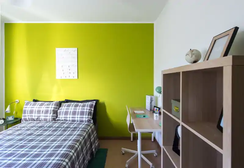 Dormitorio moderno y luminoso con cama doble frente a una pared verde intensa, ropa de cama a cuadros, lámpara de mesita y un pequeño escritorio — ideal para una estancia acogedora.