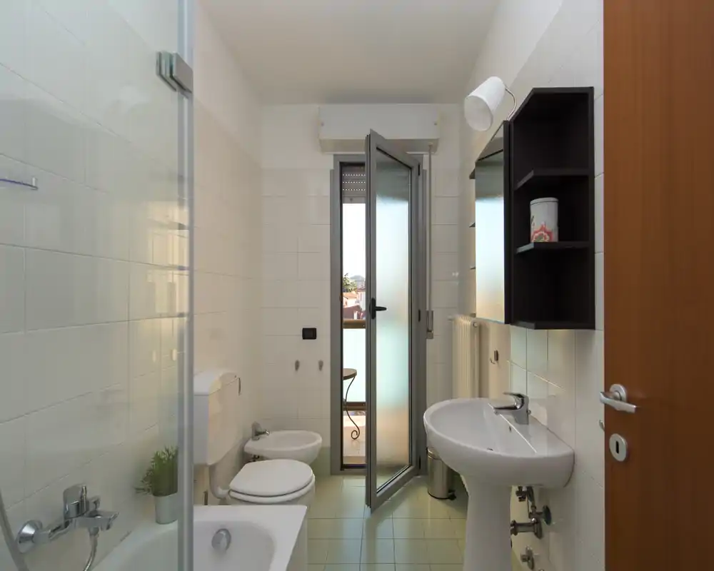 Baño limpio y luminoso con bañera, inodoro, bidé y lavabo. Un panel de ducha de cristal y una puerta a un pequeño balcón proporcionan luz natural.