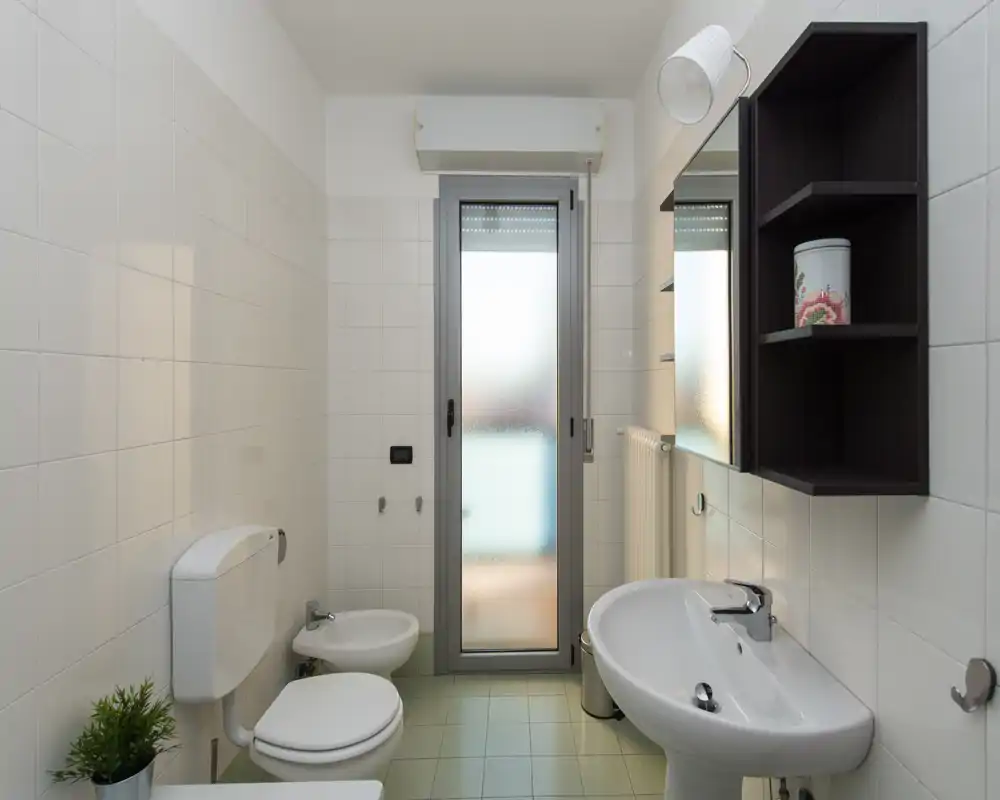 Baño limpio y luminoso con lavabo de pie, inodoro, bidé y puerta de cristal esmerilado que aporta luz natural. Paredes alicatadas en blanco y armario con espejo para un espacio funcional y ordenado.