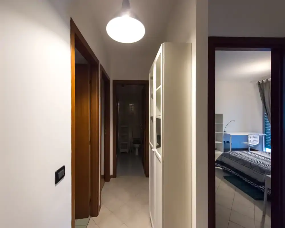 Pasillo estrecho y bien iluminado con lámpara colgante, varias puertas y vista parcial a dormitorio y baño.
