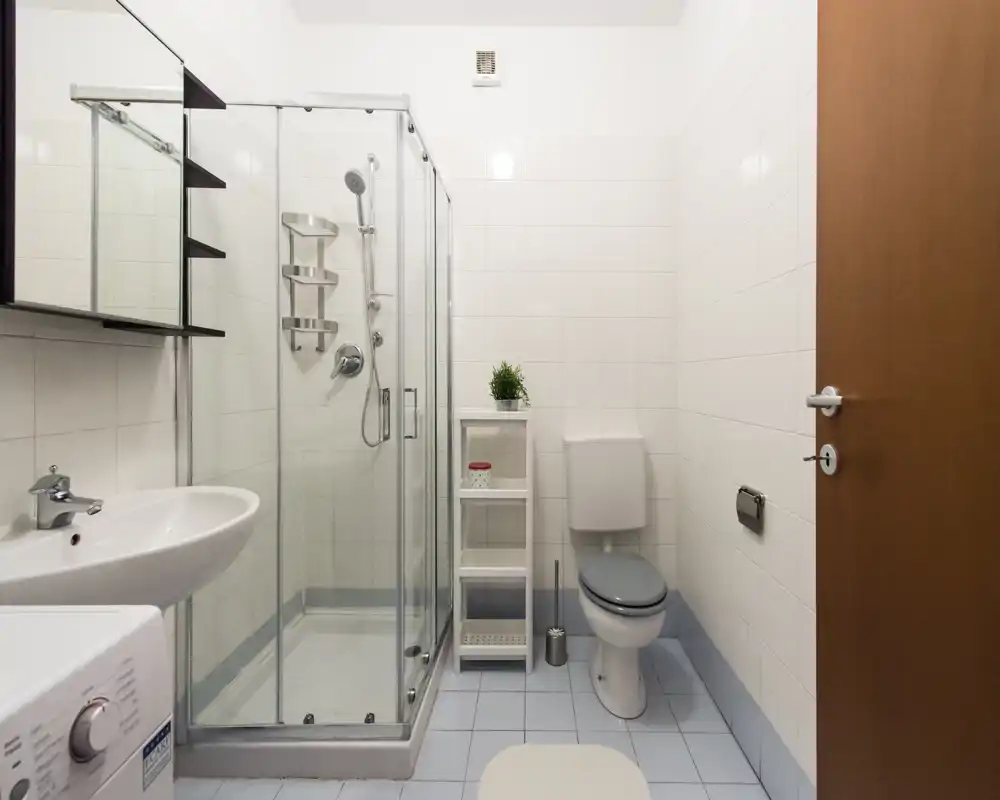 Baño compacto y limpio con ducha acristalada, inodoro, lavabo y una pequeña estantería — azulejos blancos y accesorios modernos y sencillos.
