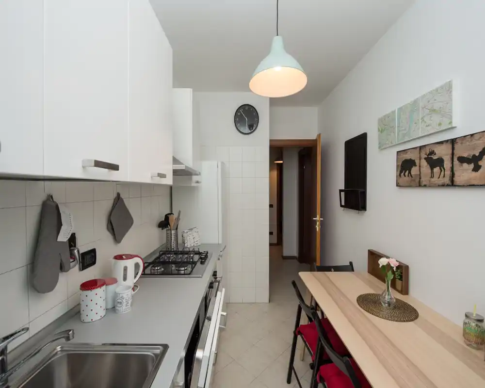 Cocina moderna y compacta con encimera larga, placa de gas y fregadero a la izquierda y una pequeña mesa con dos sillas a la derecha; tonos neutros, lámpara colgante y decoración en la pared que crean un espacio funcional y acogedor.