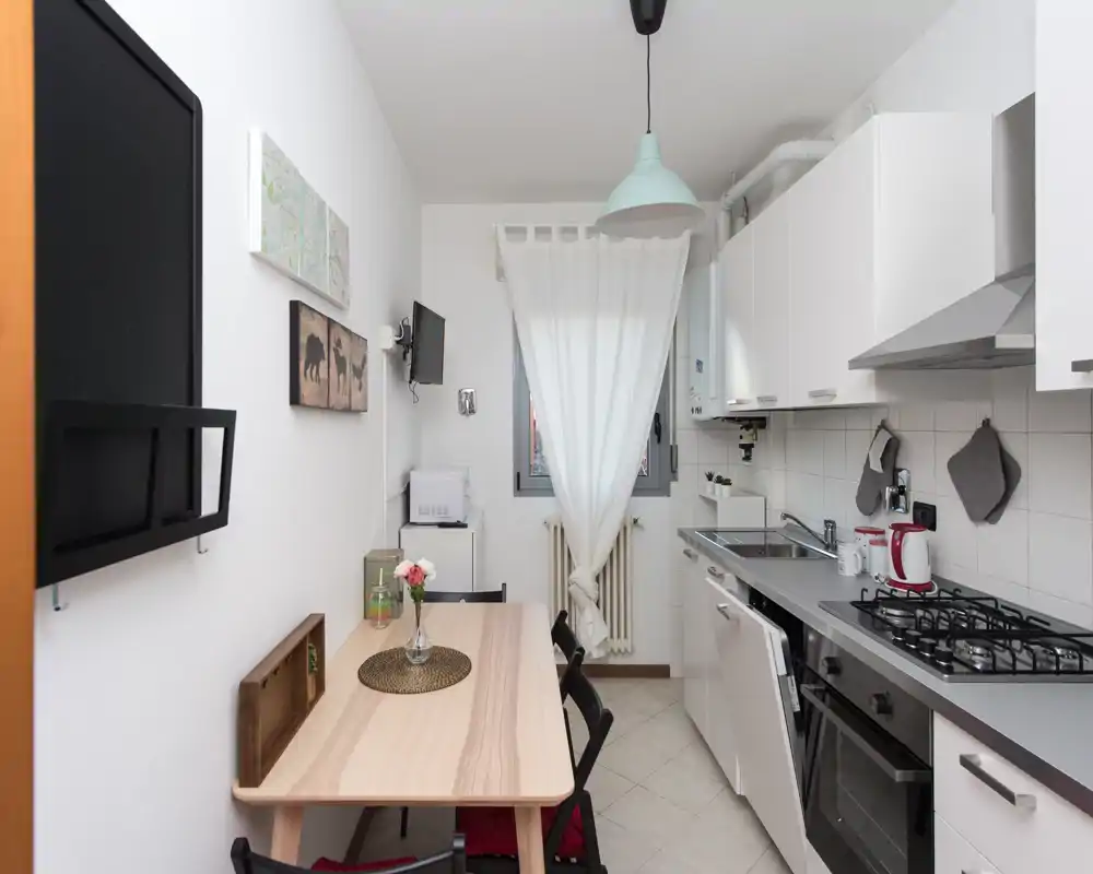 Cocina compacta y moderna con electrodomésticos integrados, mesa de madera para cuatro y luz natural suave desde la ventana con cortina — práctica y bien organizada para estancias cortas.