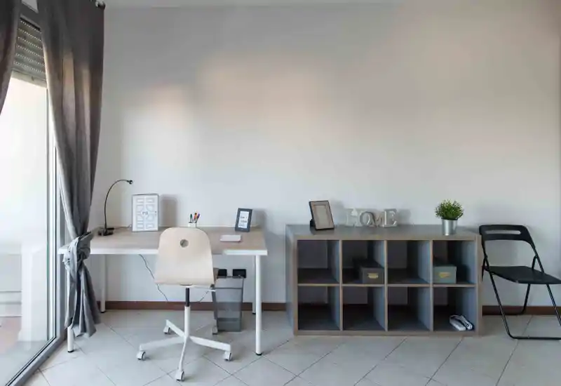Zona de estar luminosa y minimalista con escritorio ordenado y estantería modular; ideal como rincón de trabajo en casa por su luz natural y decoración moderna.