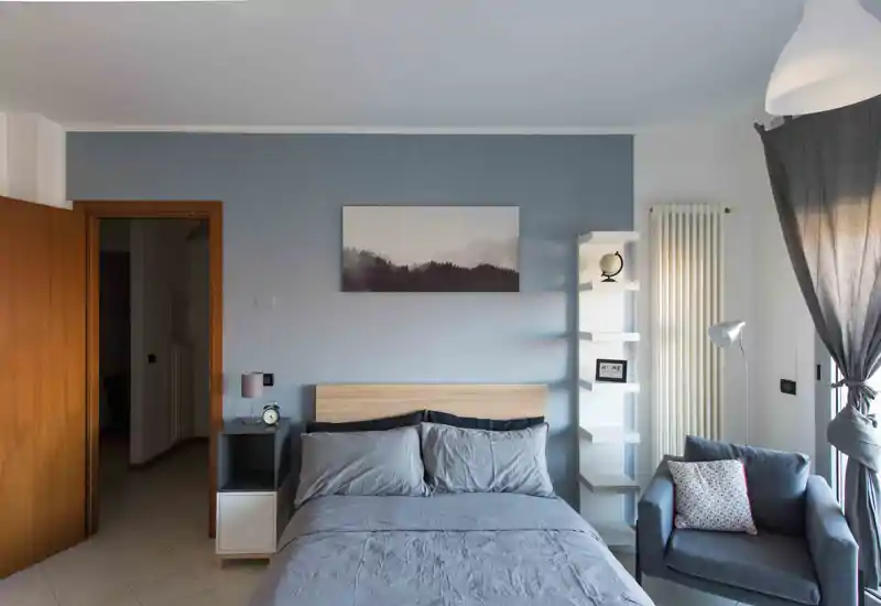 Dormitorio luminoso y ordenado con cama completa, mesita y un sillón acogedor junto a la ventana; pared en azul suave y decoración minimalista que transmite calma.