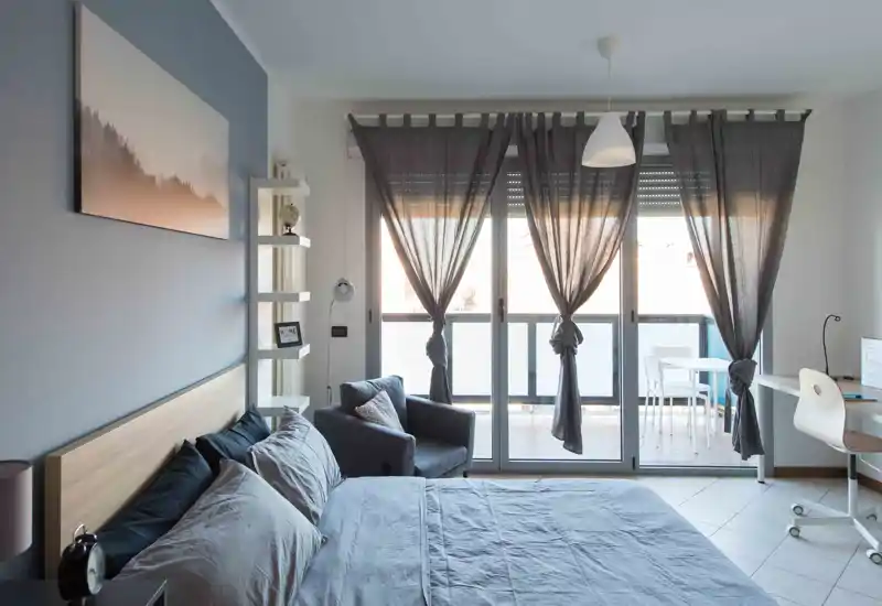 Dormitorio moderno y luminoso con una cama grande en primer plano, un sillón junto a la ventana y luz natural suave a través de las cortinas.