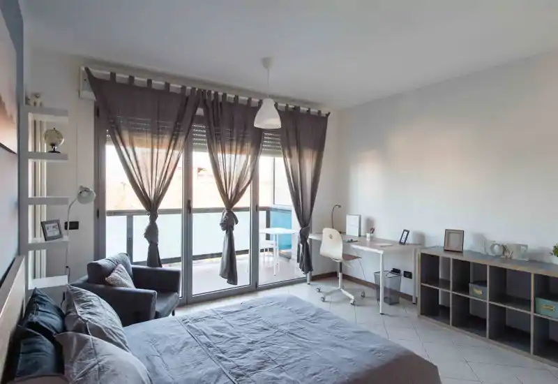 Dormitorio moderno y luminoso con una cama grande en primer plano, un sillón acogedor y un espacio de trabajo junto a las puertas correderas hacia el balcón.