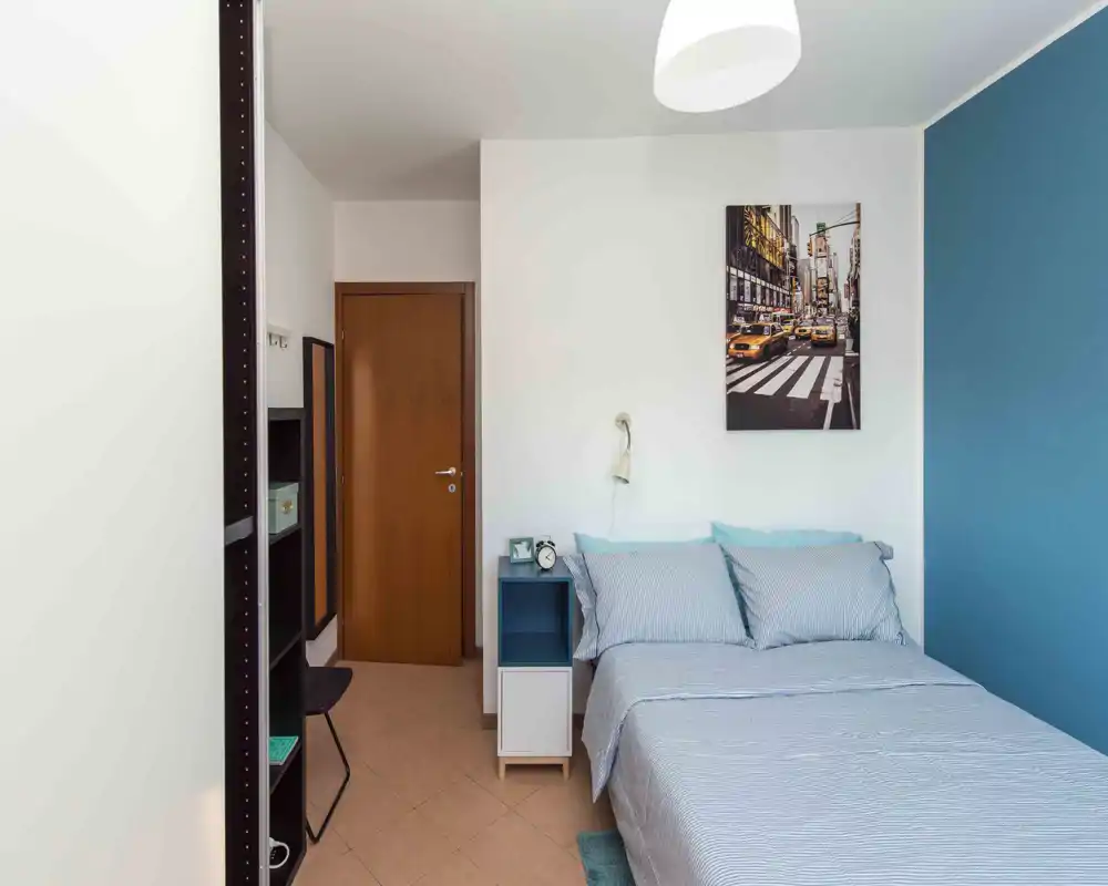 Camera da letto luminosa e ordinata con letto matrimoniale visibile, biancheria a righe, comodino e quadro a parete. Parete accentata blu e lampada moderna che creano un’atmosfera accogliente e contemporanea perfetta per annunci.