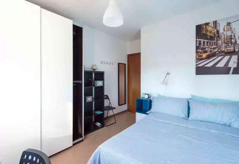 Dormitorio luminoso y ordenado con cama doble con ropa de cama azul, armario, estantería y mesita de noche — ideal para una estancia cómoda.