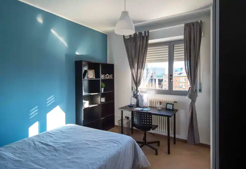 Dormitorio luminoso y ordenado con cama doble, escritorio junto a la ventana y una estantería alta frente a una pared color turquesa — perfecto para trabajar y descansar con abundante luz natural.