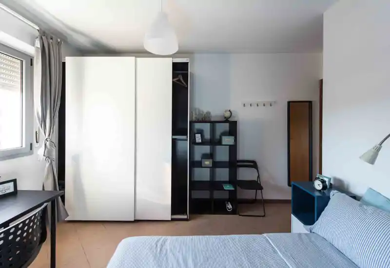 Dormitorio moderno y luminoso con cama doble bien tendida, estantería junto a la mesilla y un gran armario. La luz natural de la ventana y la decoración minimalista crean un ambiente tranquilo, perfecto para un anuncio.