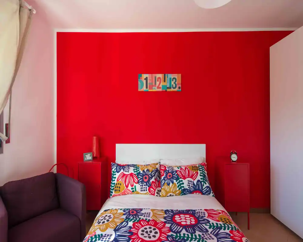 Chambre lumineuse et colorée avec un mur rouge vif, coussins à motifs et une tête de lit blanche épurée — chaleureuse et bien mise en scène pour une annonce.