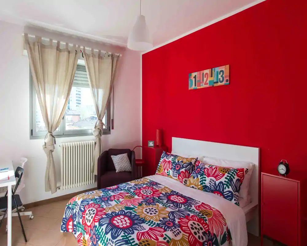 Chambre lumineuse et colorée avec un mur d'accent rouge et une literie à motifs. La lumière naturelle de la fenêtre met en valeur un fauteuil cosy et des détails de chevet minimalistes.