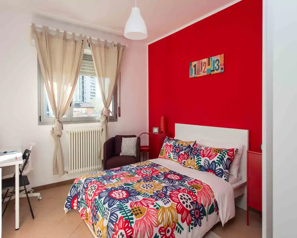 Chambre lumineuse et colorée avec un lit double aux couettes florales vives, un mur d'accent rouge, un petit fauteuil et un bureau près de la fenêtre — idéale pour une annonce chaleureuse et stylée.