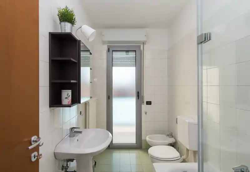 Bright, clean bathroom with a pedestal sink, bidet, toilet and a glass shower door; frosted window provides privacy and natural light.