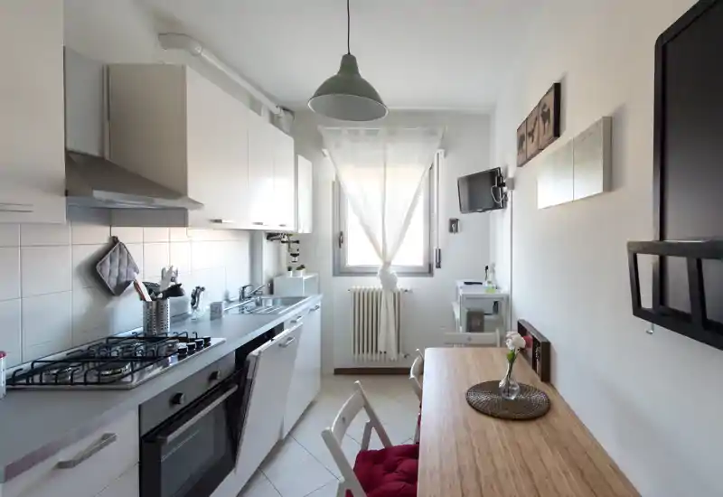 Bright, compact kitchen with integrated stove, oven and sink along one wall, a small wooden breakfast bar and seating — well organized and ideal for a city apartment.