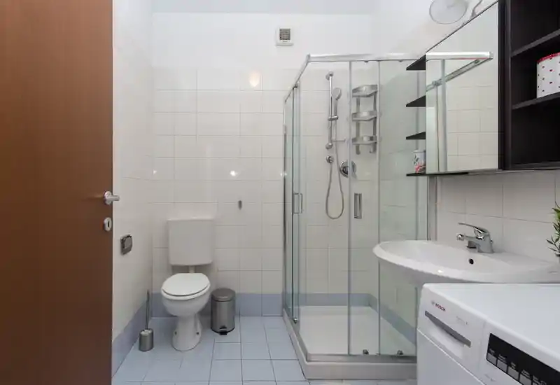 Clean modern bathroom with glass-enclosed shower, wall-mounted sink, toilet and washing machine visible; neutral tiles and bright lighting provide a fresh feel.