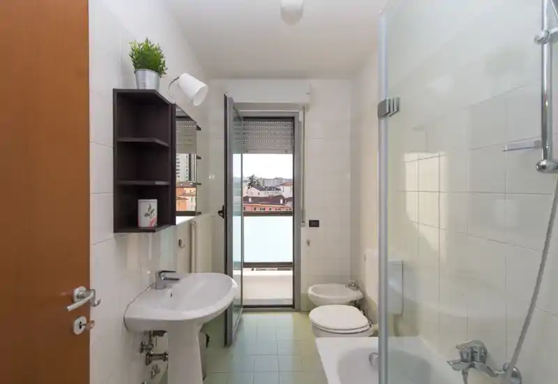 Bright, clean bathroom with a sink, toilet, bidet and bathtub with glass shower screen; balcony door provides natural light and a city view.
