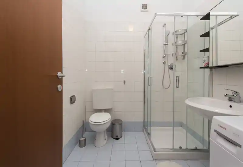 Clean, modern bathroom with tiled walls, a corner glass shower, toilet, sink and compact storage. Bright neutral lighting and minimalist fittings make it practical and easy to maintain.