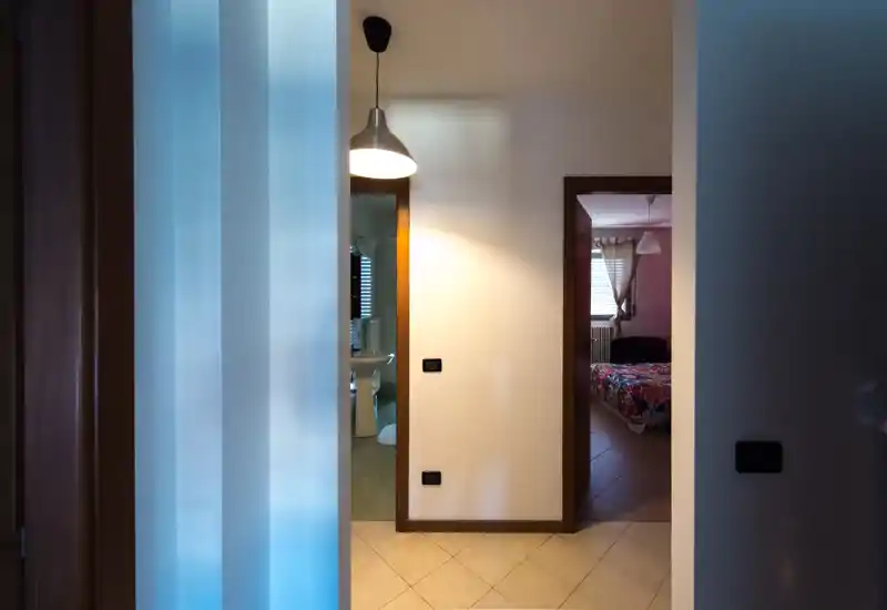 Hallway view leading to a bedroom: a partially visible bed through the open doorway with patterned bedding, neutral tiled floor and soft pendant lighting in the corridor.