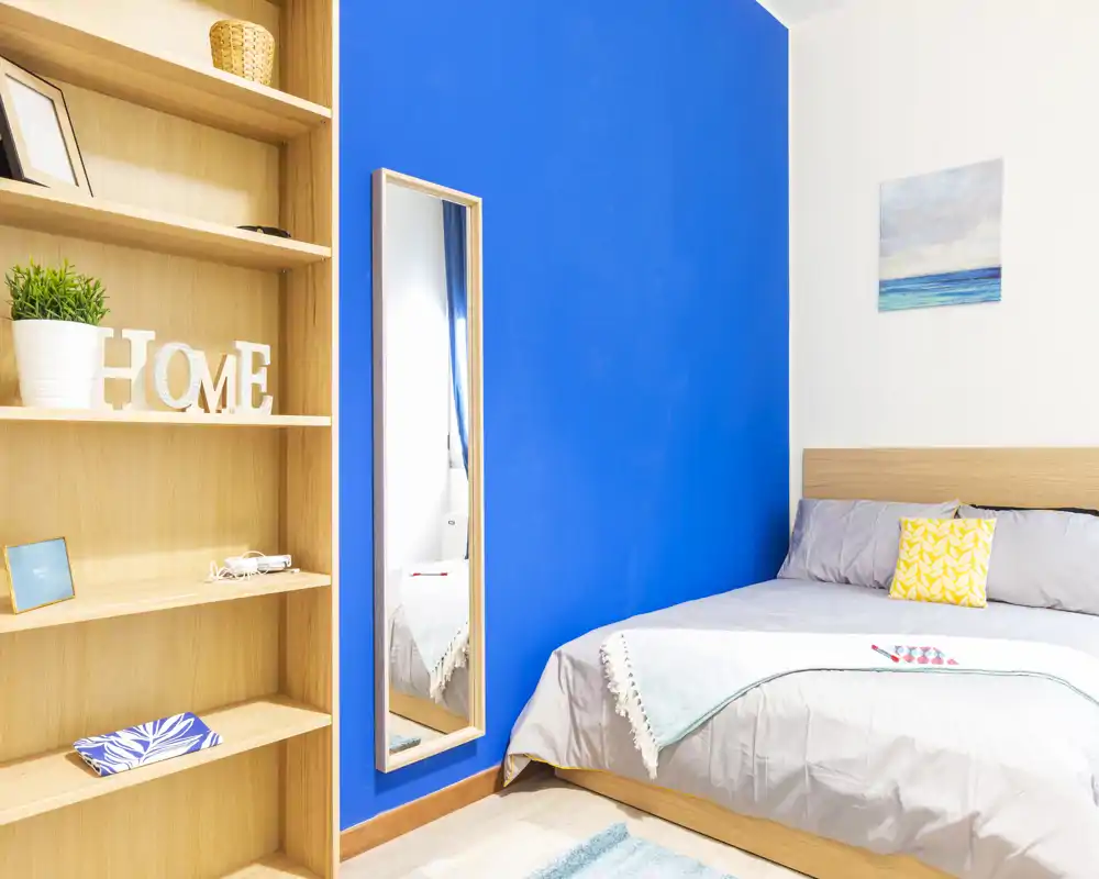 Bright, contemporary bedroom with a vivid blue accent wall, a neatly made double bed, decorative cushions and a full-length mirror. Clean, modern shelving and soft textiles create a welcoming atmosphere for short-term rentals.