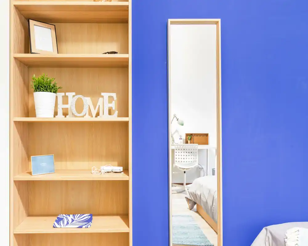 Bright, modern bedroom corner with a wooden shelf and a tall mirror reflecting a tidy bed and desk area; decorative details include a potted plant and 'HOME' letters for a welcoming feel.