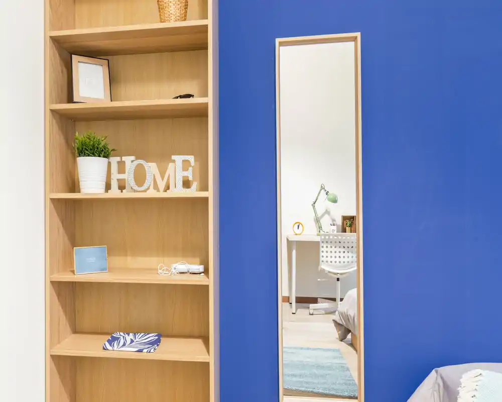 Compact bedroom corner with a blue accent wall, open wooden shelving and a narrow full-length mirror reflecting a bedside desk; bright, tidy and simply styled.