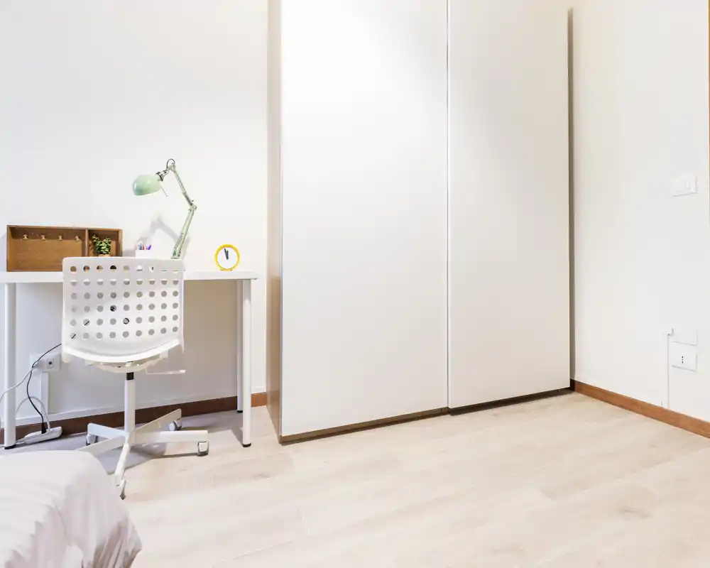 Minimal, bright bedroom corner with a small white desk, perforated chair, bedside area and a large white wardrobe. Clean, modern styling and ample natural-like light make the space feel airy and functional.